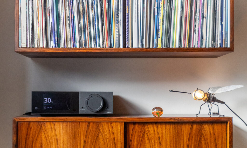 Enjoy the Music reviews the Lyngdorf TDAI-2210 10 Lyngdorf TDAI-2210 streaming integrated amplifier with RoomPerfect™ room correction displayed on a wooden cabinet beneath a vinyl record collection in a modern listening room.